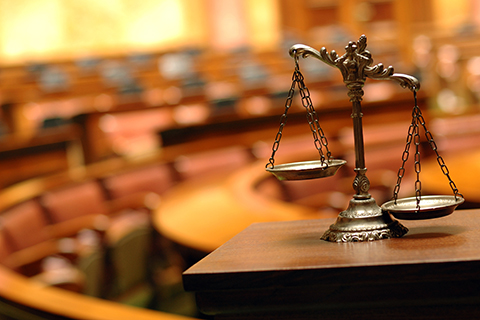 This is a stock photo from Shutterstock. In the foreground are the "scales of justice." The background is blurred. Court room seating is in the background. 